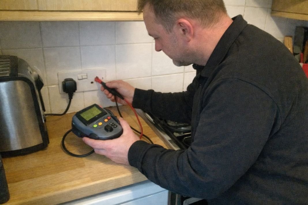 An illustrative image of an engineer testing electrical UK plug sockets with a portable device in a compact kitchen